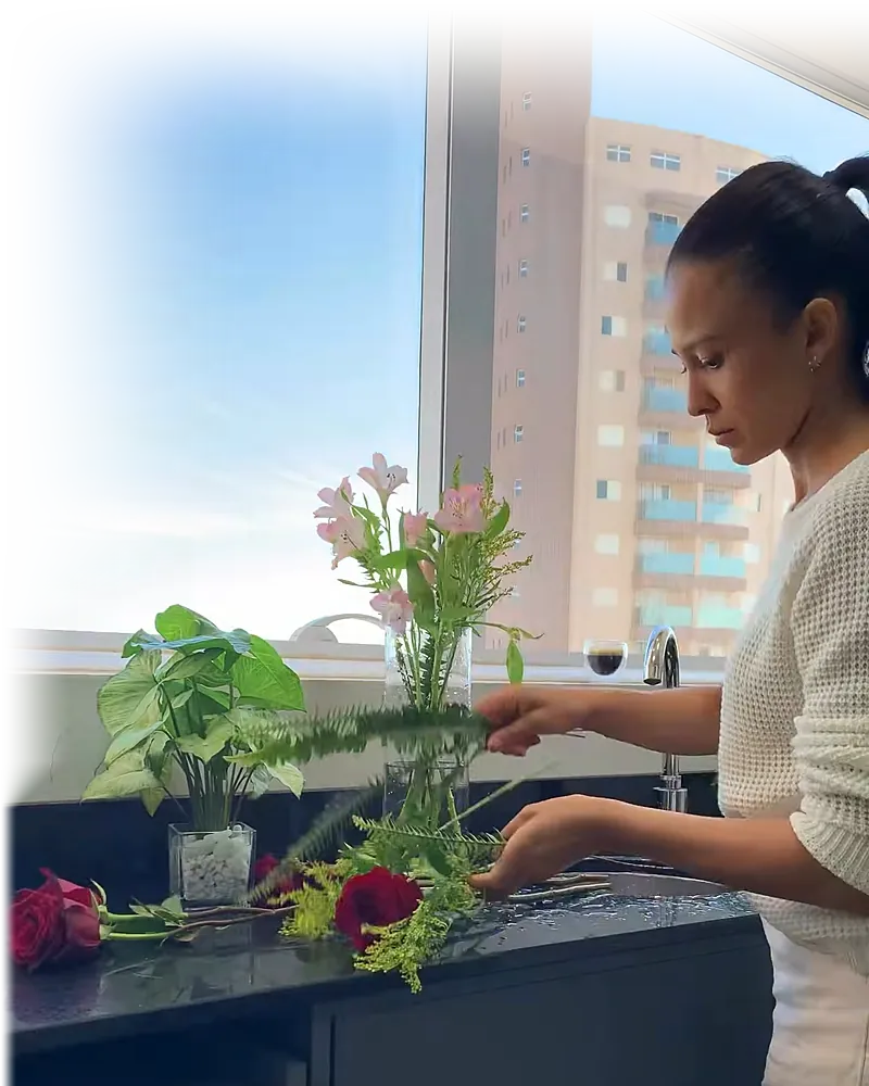 Dra. Francielle, de perfil, apara flores na bancada de granito da sala clínica; pela janela, o céu aberto e um prédio da cidade.