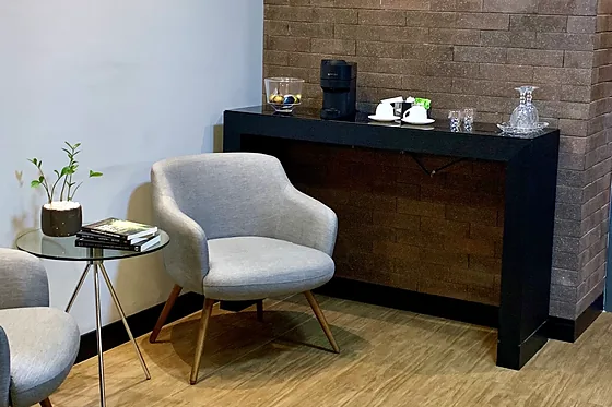 Recepção do consultório: poltronas cinza de pé palito, mesinha de vidro com uma planta e o aparador do café diante da parede de tijolinho.