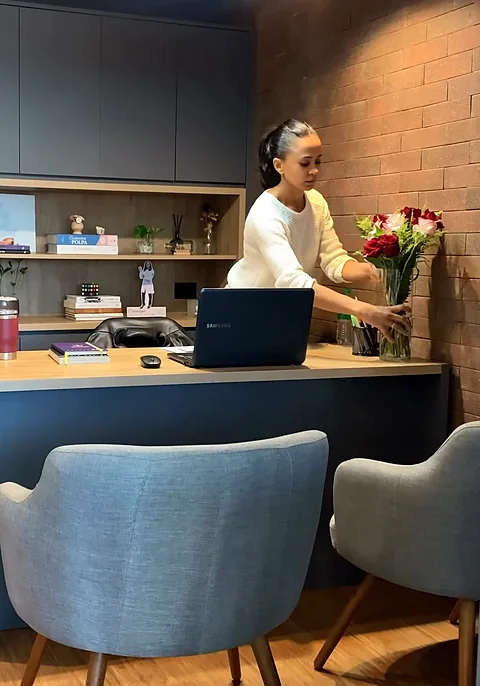 Dra. Francielle põe um vaso de flores sobre a mesa de carvalho da sala de conversa; atrás, a marcenaria grafite e a parede de tijolinho.