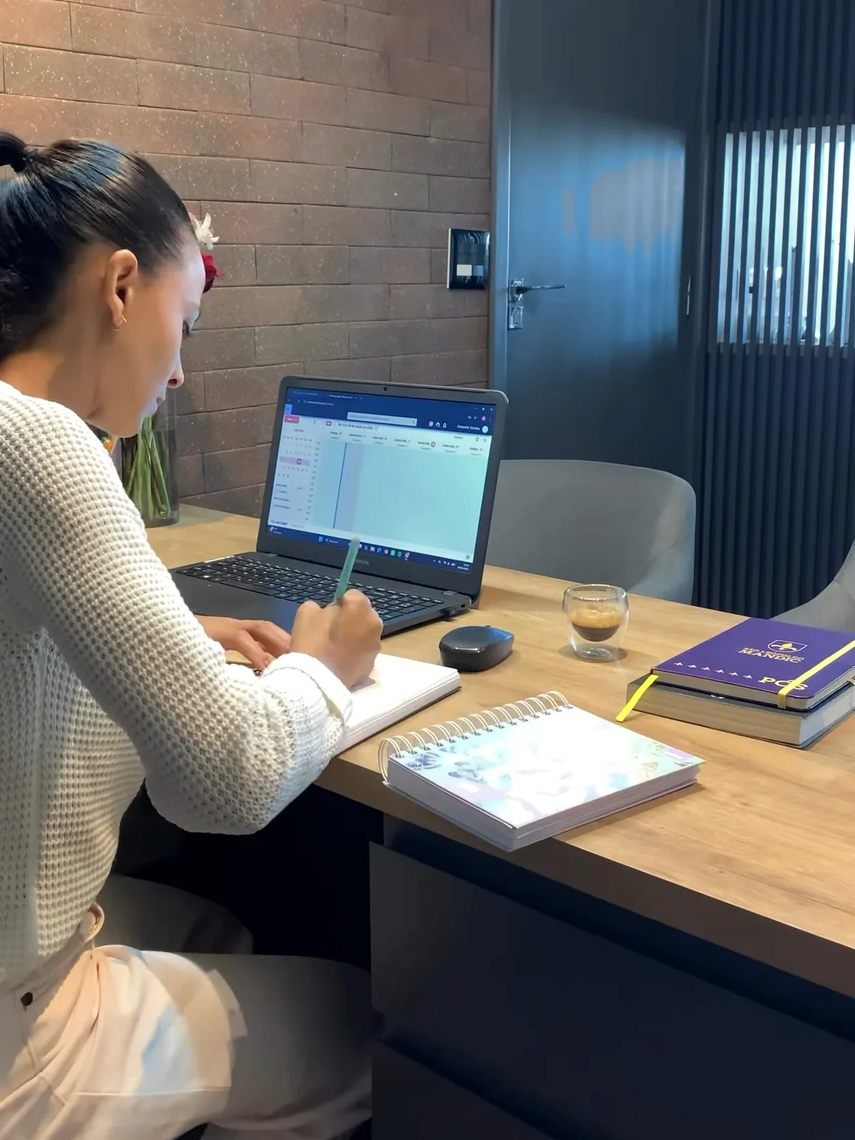 A Dra. Francielle, vista de lado, escreve num caderno sobre a mesa de carvalho. Ao lado dela, o notebook com a agenda aberta, um café e a agenda de espiral; do outro lado da mesa, uma poltrona cinza vazia.